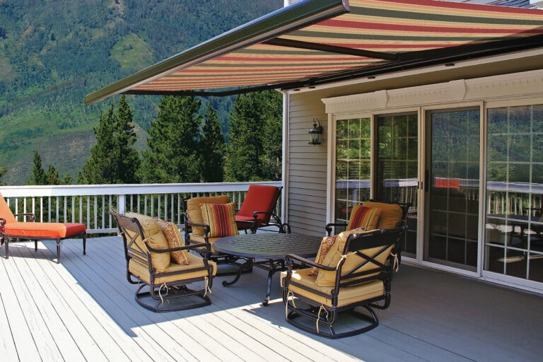 Outdoor deck with cushioned patio furniture under a striped retractable awning, overlooking forested mountains.