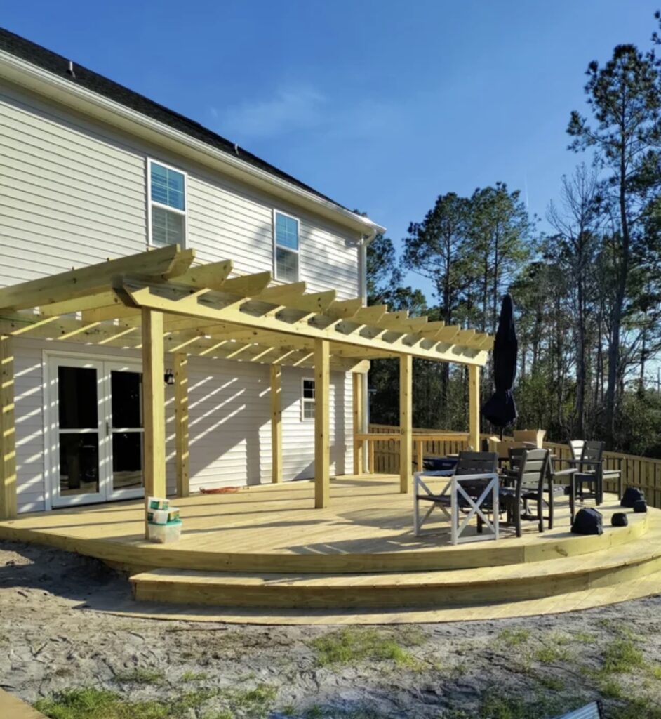 Newly built wooden deck with pergola attached to a house, featuring outdoor dining furniture and surrounded by trees.