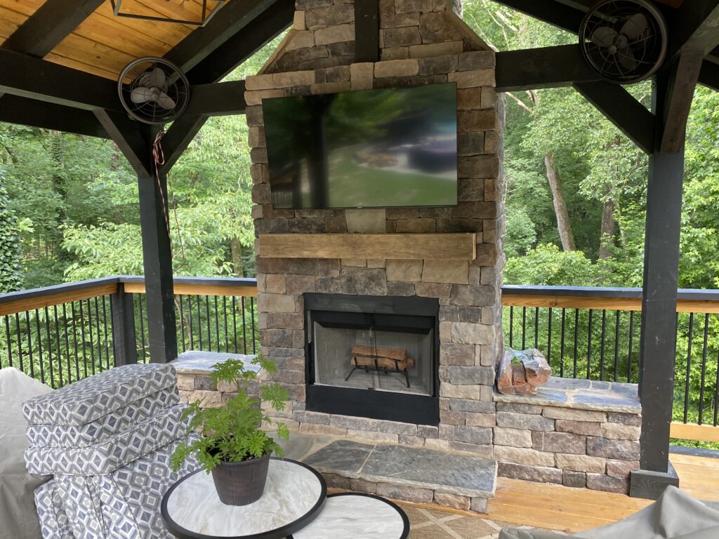 Outdoor covered patio with a stacked stone fireplace, mounted TV above, ceiling fans, cushioned seating, potted plant on round table